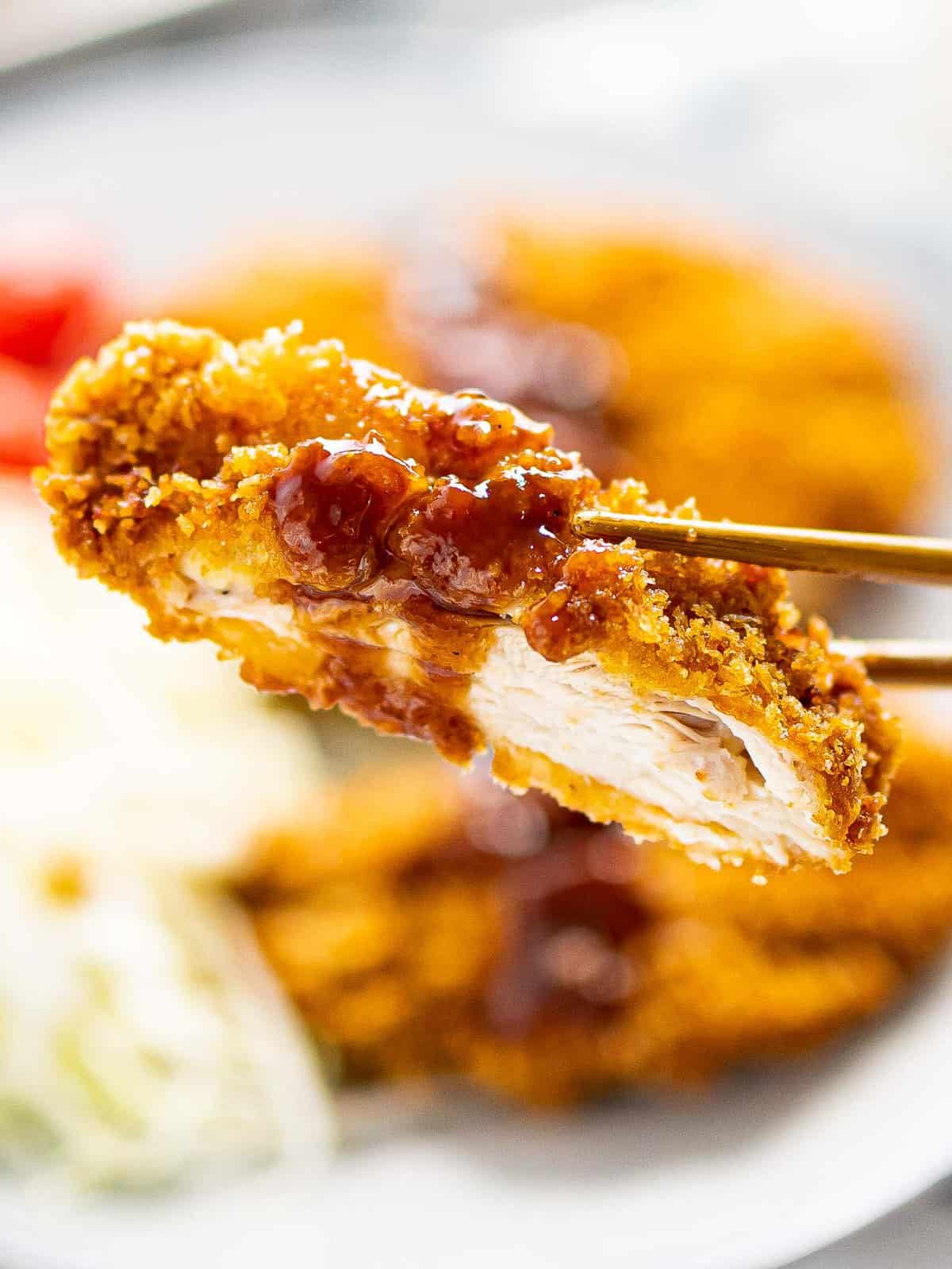 Close up of a piece of chicken katsu with katsu sauce held by chopsticks.